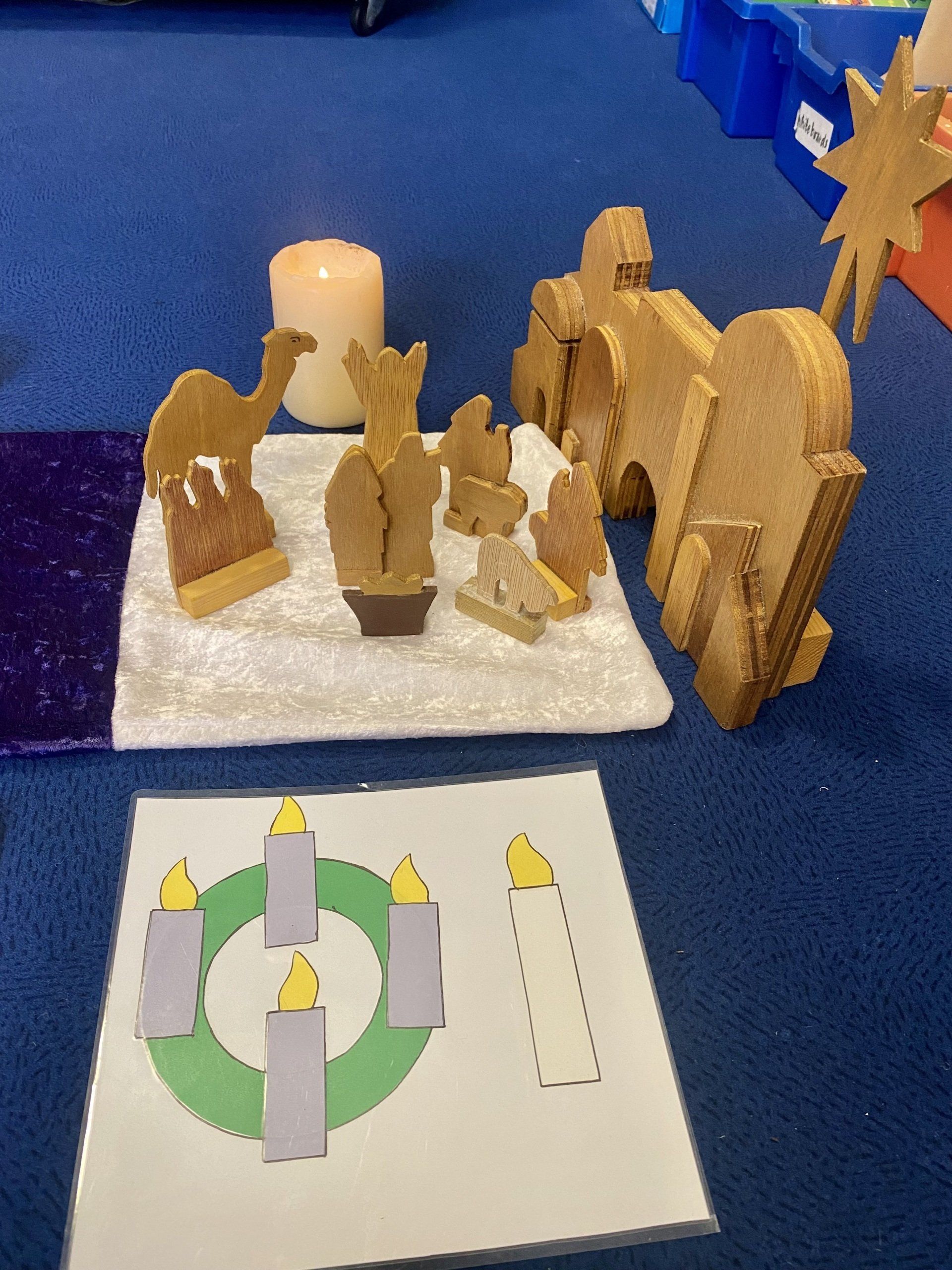 Nativity scene with wooden figures, candle, and Advent wreath image on blue carpet.
