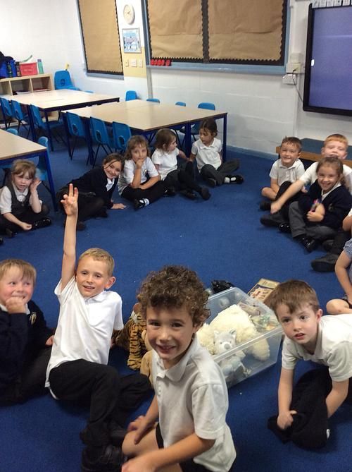 Children in a classroom, sitting in a circle on the floor. One boy raises his hand.