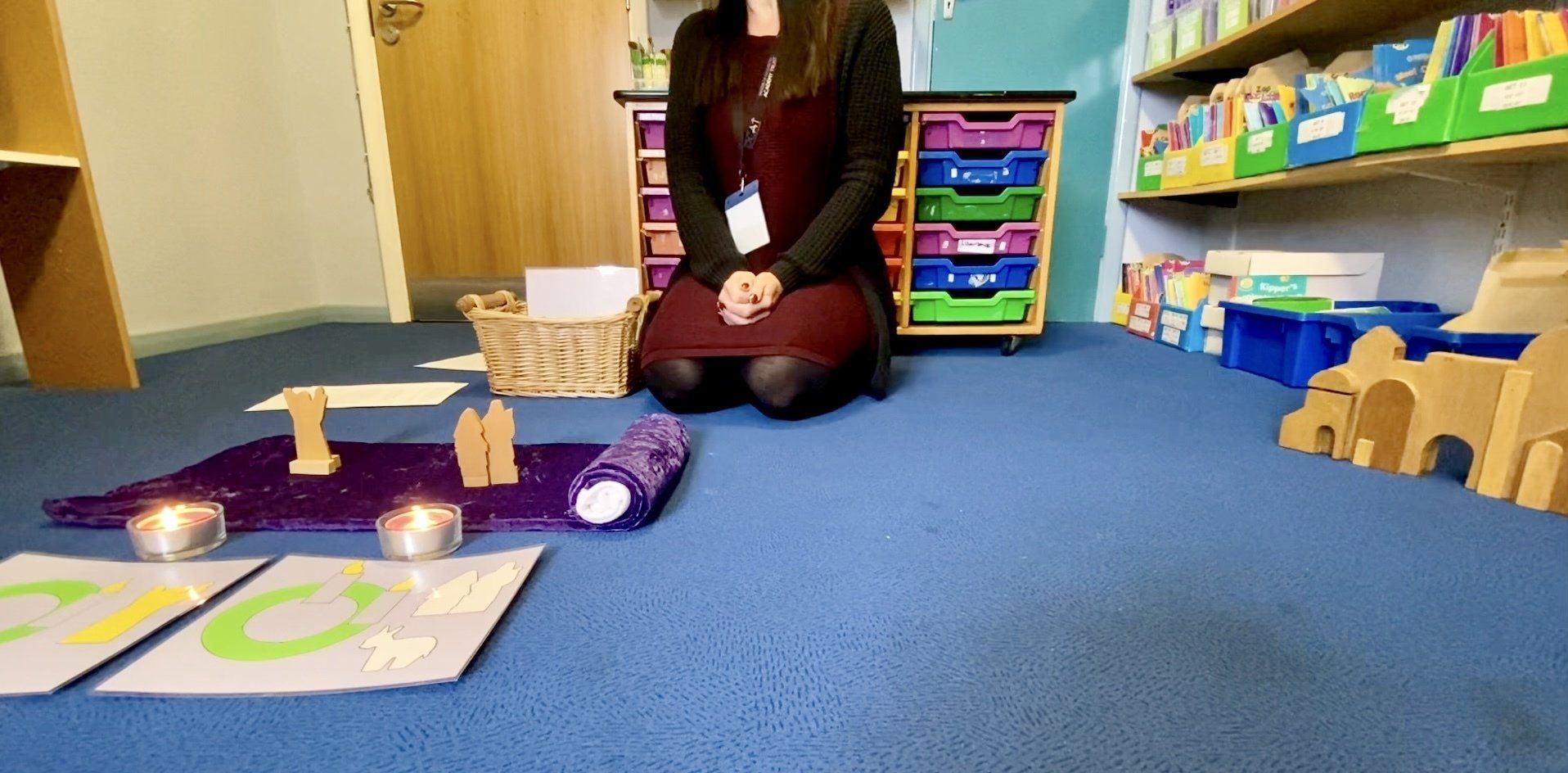 Person kneeling on a blue carpet with candles, purple cloth, and wooden objects. Shelves and drawers in the background.