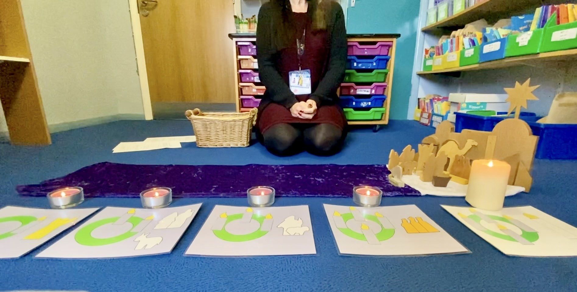 A person kneels before candles and art; a purple cloth and a Nativity scene on the floor in a classroom.