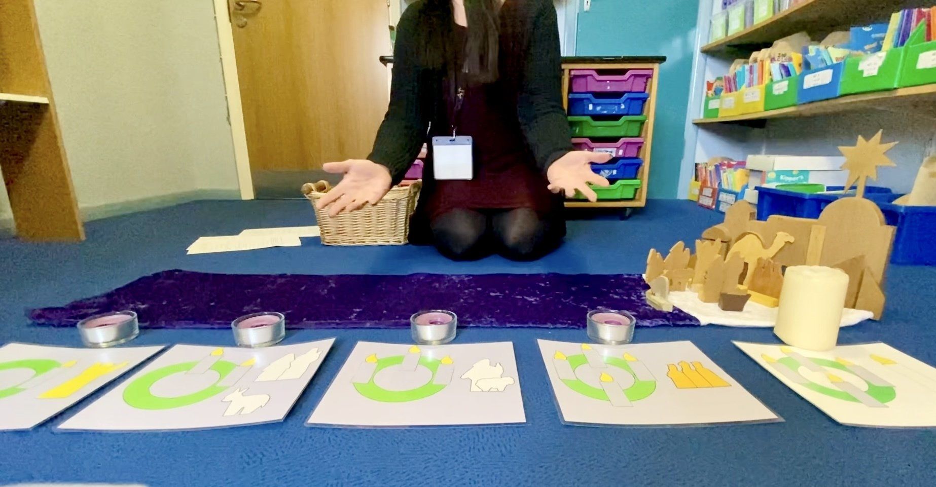 Person kneeling, gesturing toward Advent display: purple cloth, candles, drawings of wreaths, and nativity scene.