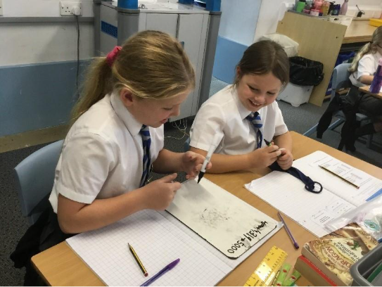 Two students in white shirts and ties work on a math problem at a desk, smiling.