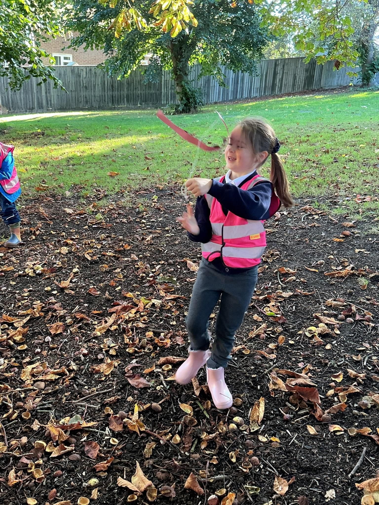 Child in a pink vest swings a stick in a leaf-covered yard near a wooden fence.