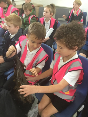 Children in pink safety vests sit in classroom chairs, looking at a black bag or costume piece.