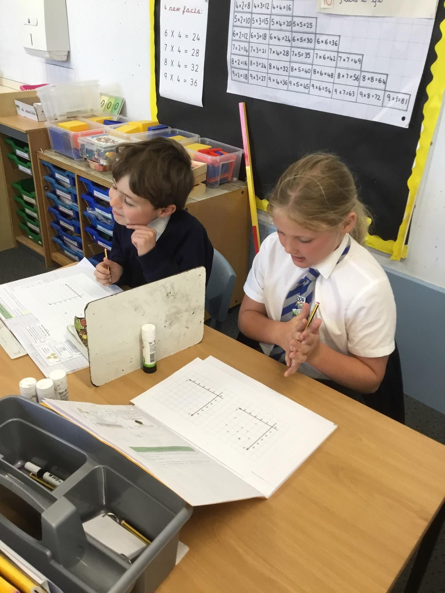Two students working at a classroom table with papers and a laptop; one uses a pen, the other adjusts a tie.