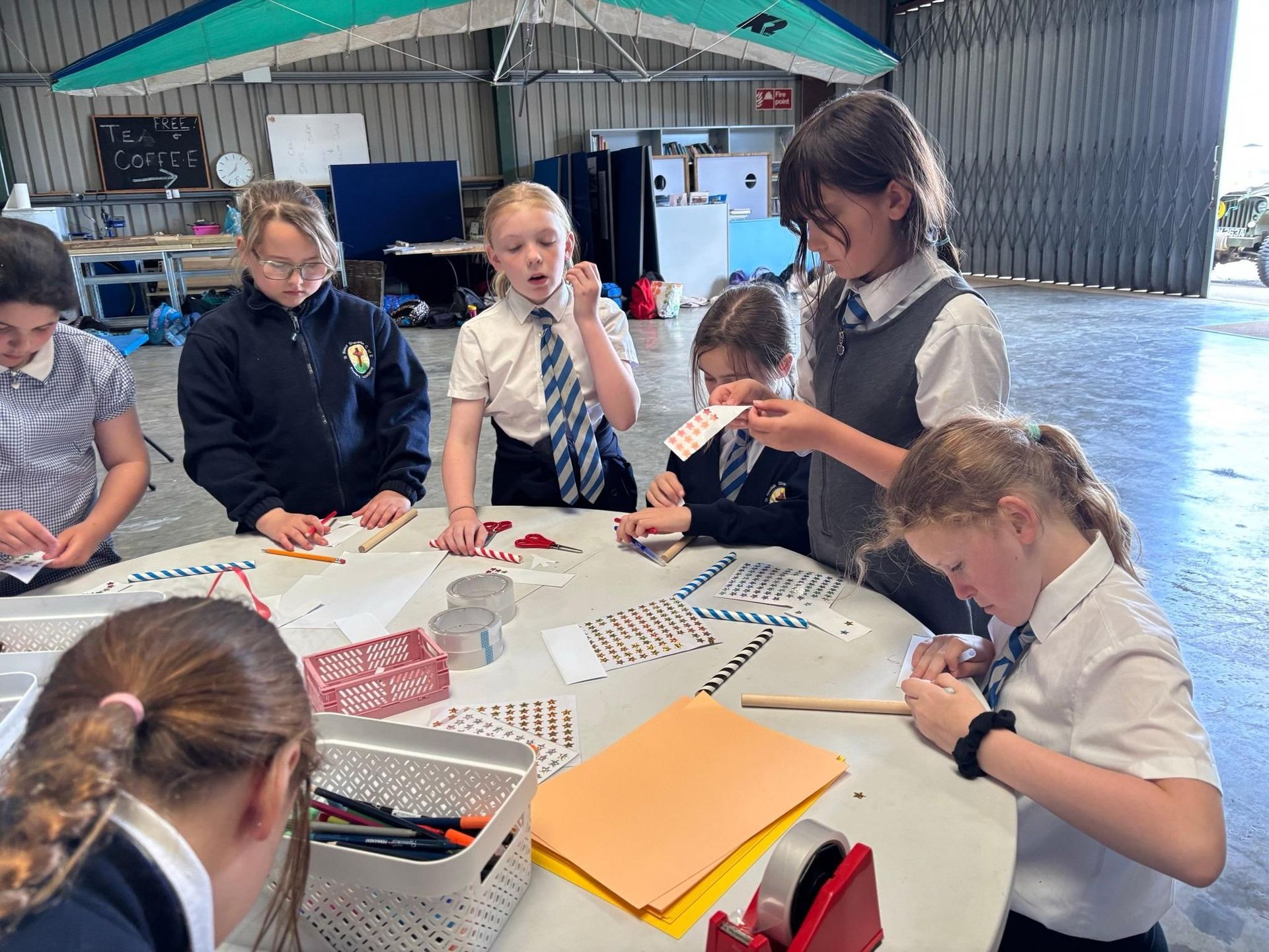 Children doing a hands-on craft activity at a table in a workshop or classroom, with paper, markers, and notebooks.