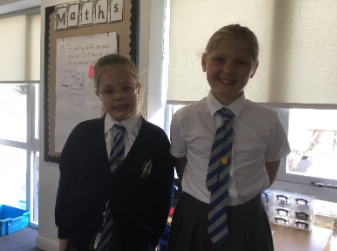 Two children in school uniforms standing in a classroom, smiling at the camera.