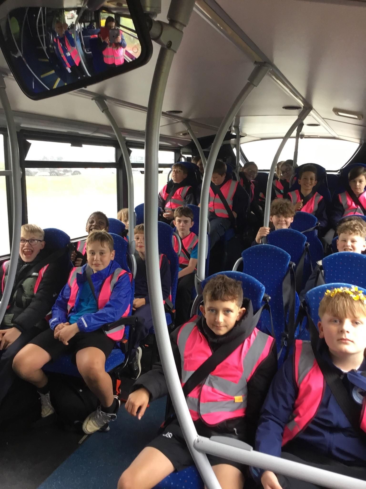 Children seated on a bus, wearing blue jackets and pink safety vests, reflected in the rearview mirror.