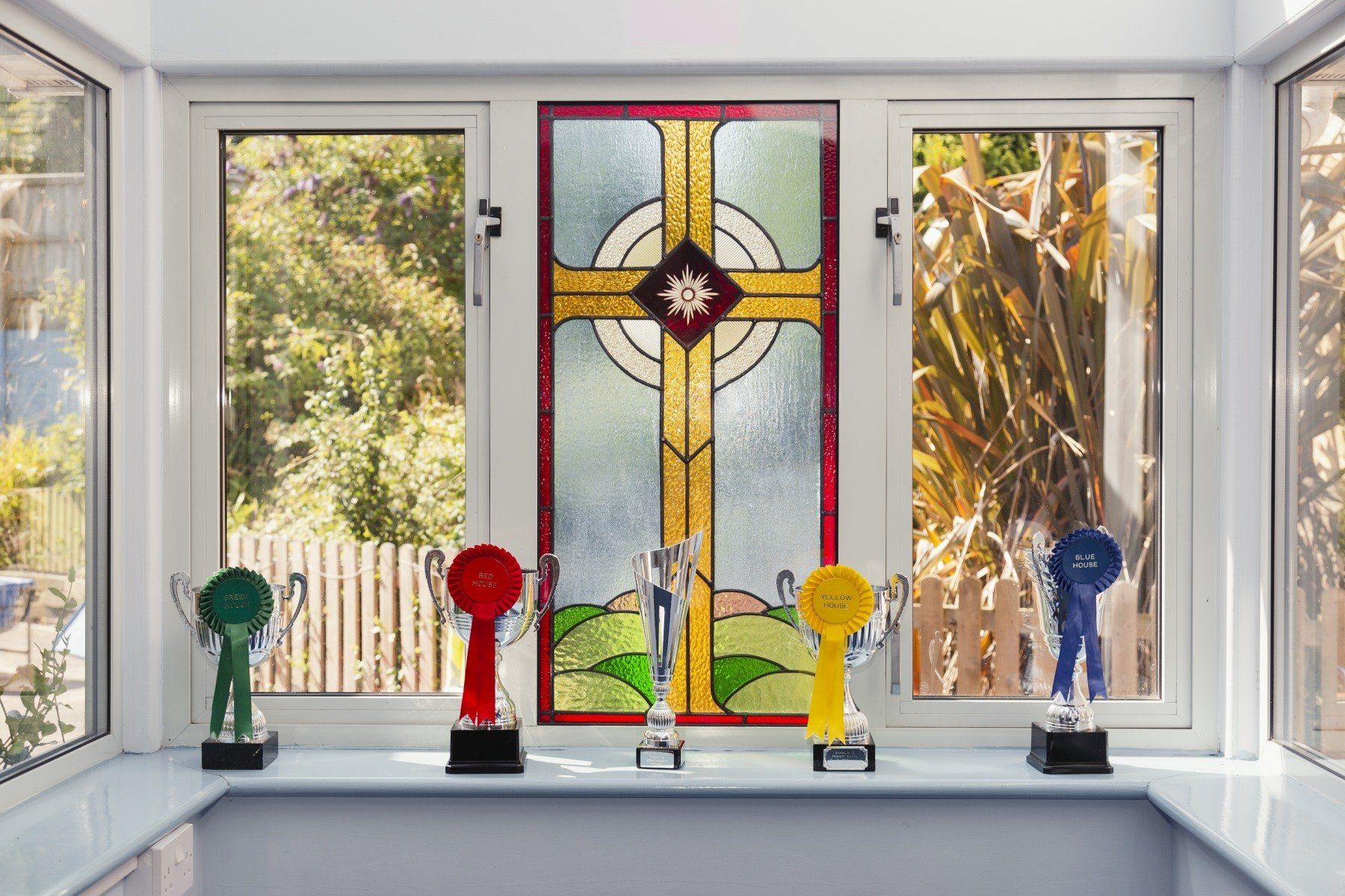 Window with colorful ribbons and trophies, stained glass cross in center, outside greenery.