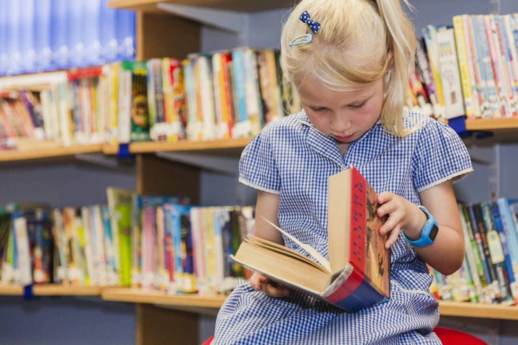 Girl in school uniform reading a book in a library, surrounded by bookshelves.