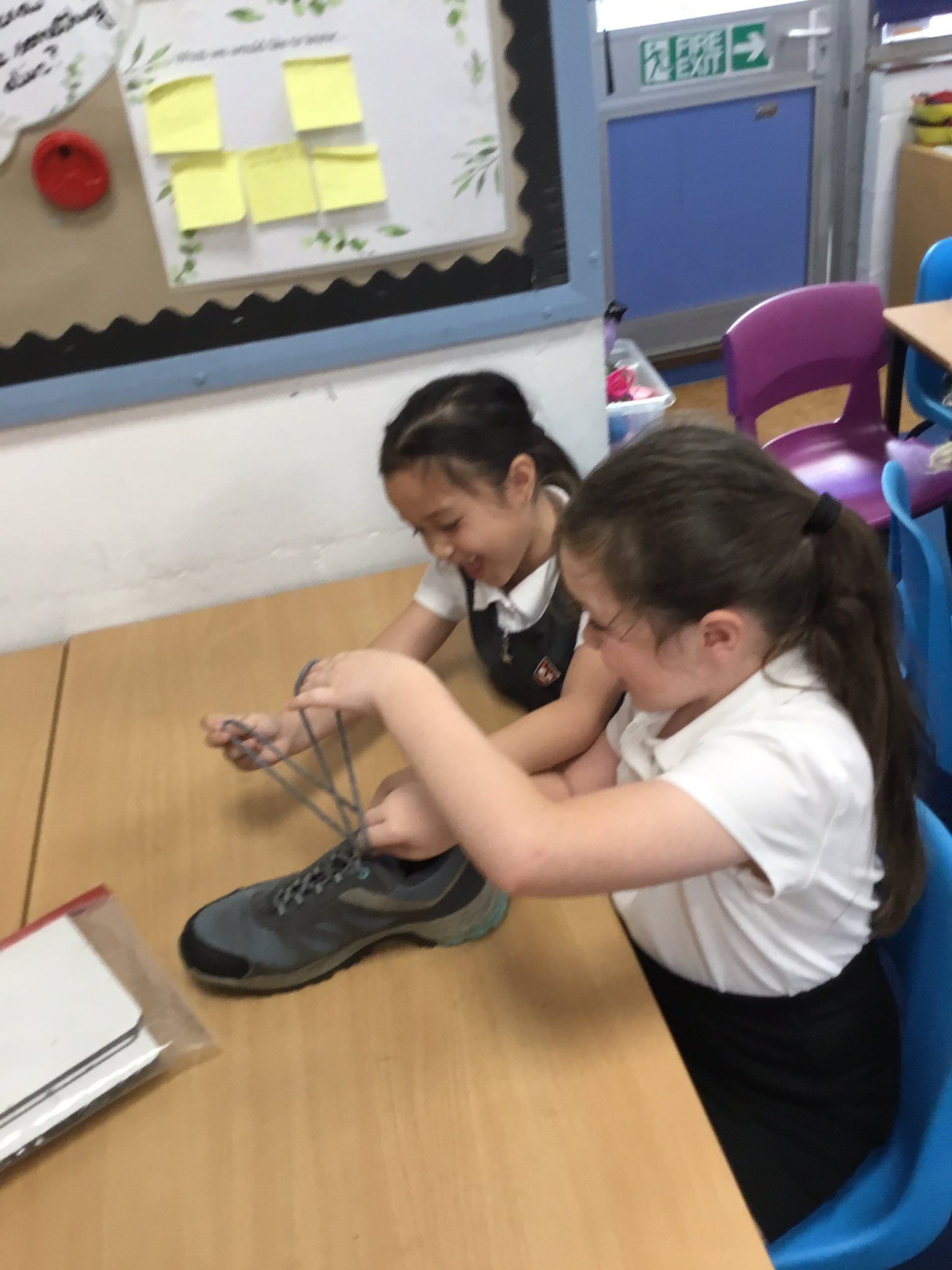 Two young girls, at a desk, one tying a shoe, other watches. Classroom setting.