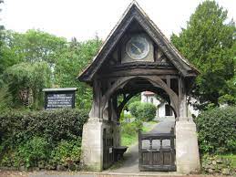 Wooden lychgate with clock and open gate leading to a church. A sign is to the left.