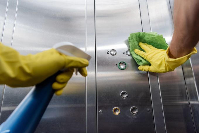 Mani con guanti gialli disinfettano i pulsanti in acciaio inossidabile dell'ascensore con un panno verde.