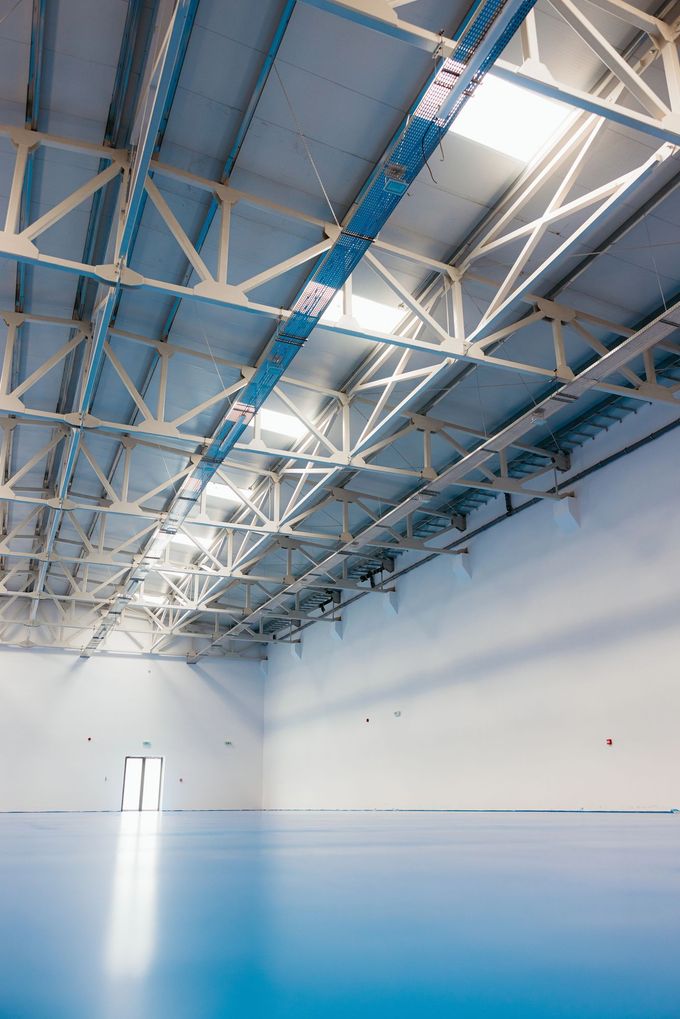 Magazzino interno vuoto con pavimento blu, pareti bianche e soffitto alto con travi metalliche e lucernari.