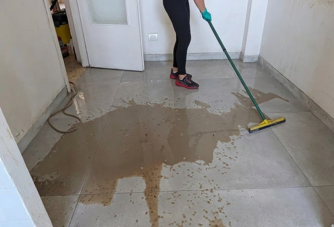 Una persona lava via l'acqua fangosa dal pavimento piastrellato di un corridoio.