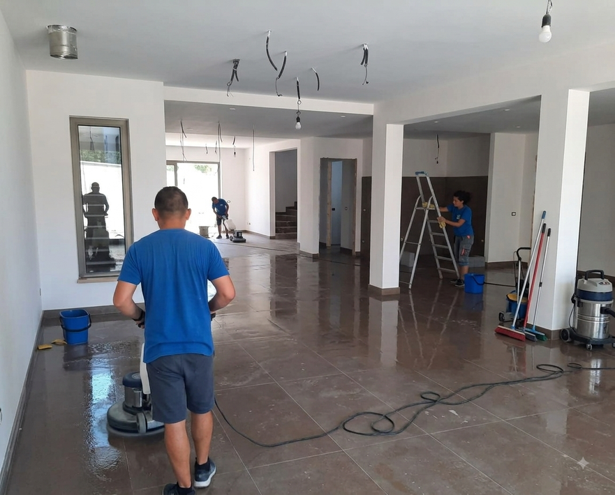 Operaio che pulisce una stanza luminosa e vuota, con attrezzi e cavi sul pavimento, in un cantiere o in fase di ristrutturazione.