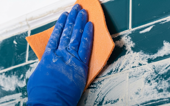 Una mano con un guanto blu strofina una parete piastrellata color verde acqua con una spugna arancione.