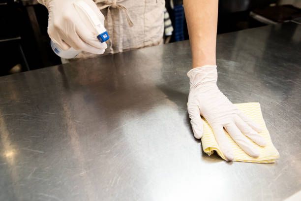 Mani guantate che puliscono un piano di lavoro in acciaio inossidabile con un panno e un flacone spray.