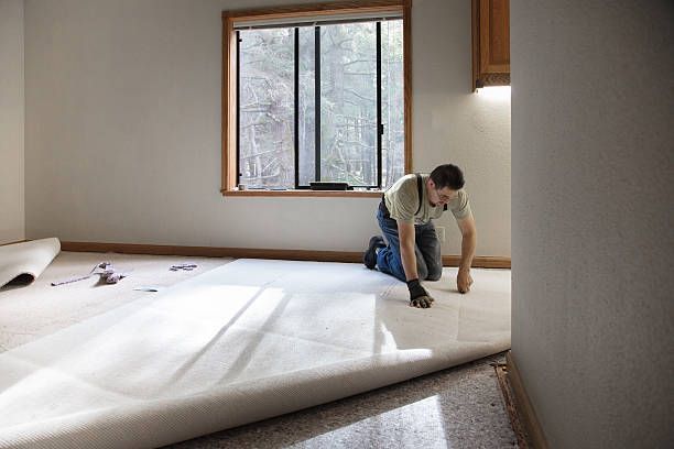 Person kneeling on a bare floor, measuring or smoothing white flooring material in a room with a window.