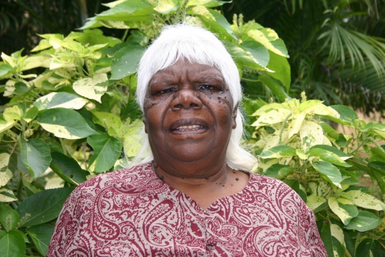 A woman with white hair is standing in front of a bush.