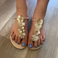 Feet in beige sandals with white floral straps and blue toenail polish on a wooden floor