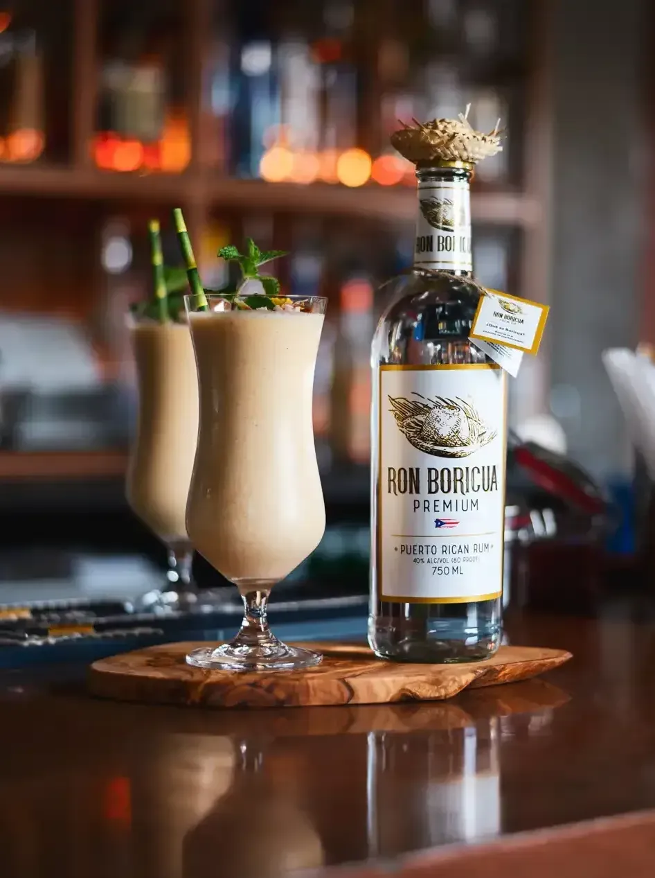 A bottle of Ron Boricua is sitting on a wooden table next to two glasses of Puerto Rican Piña Colada Drink.