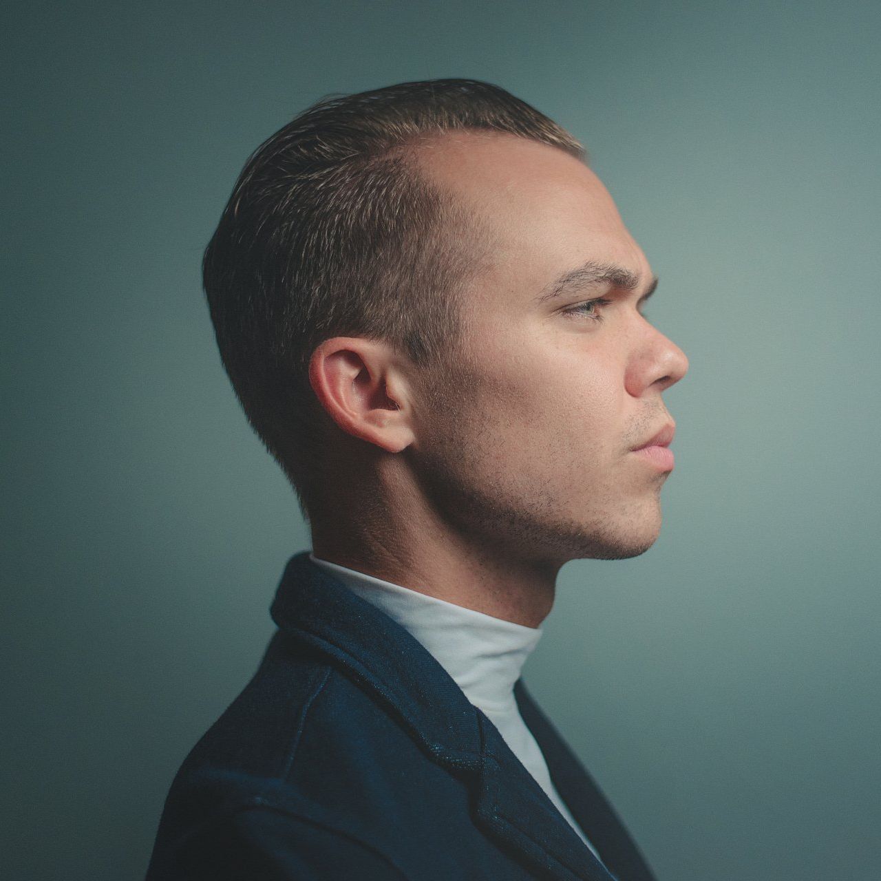 A man in a suit and white turtleneck is looking to the side.