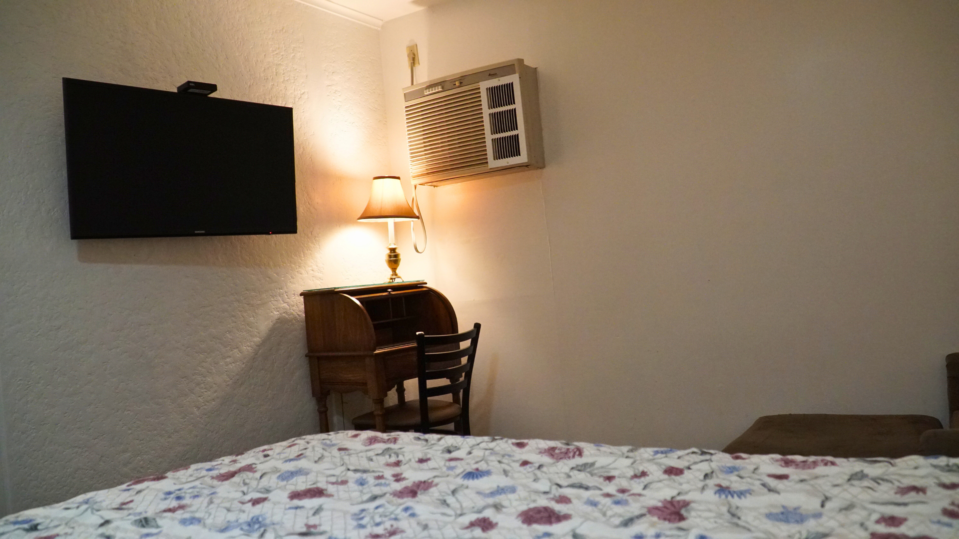 Cozy bedroom with TV, lamp-lit desk, chair, wall art, and floral bedspread