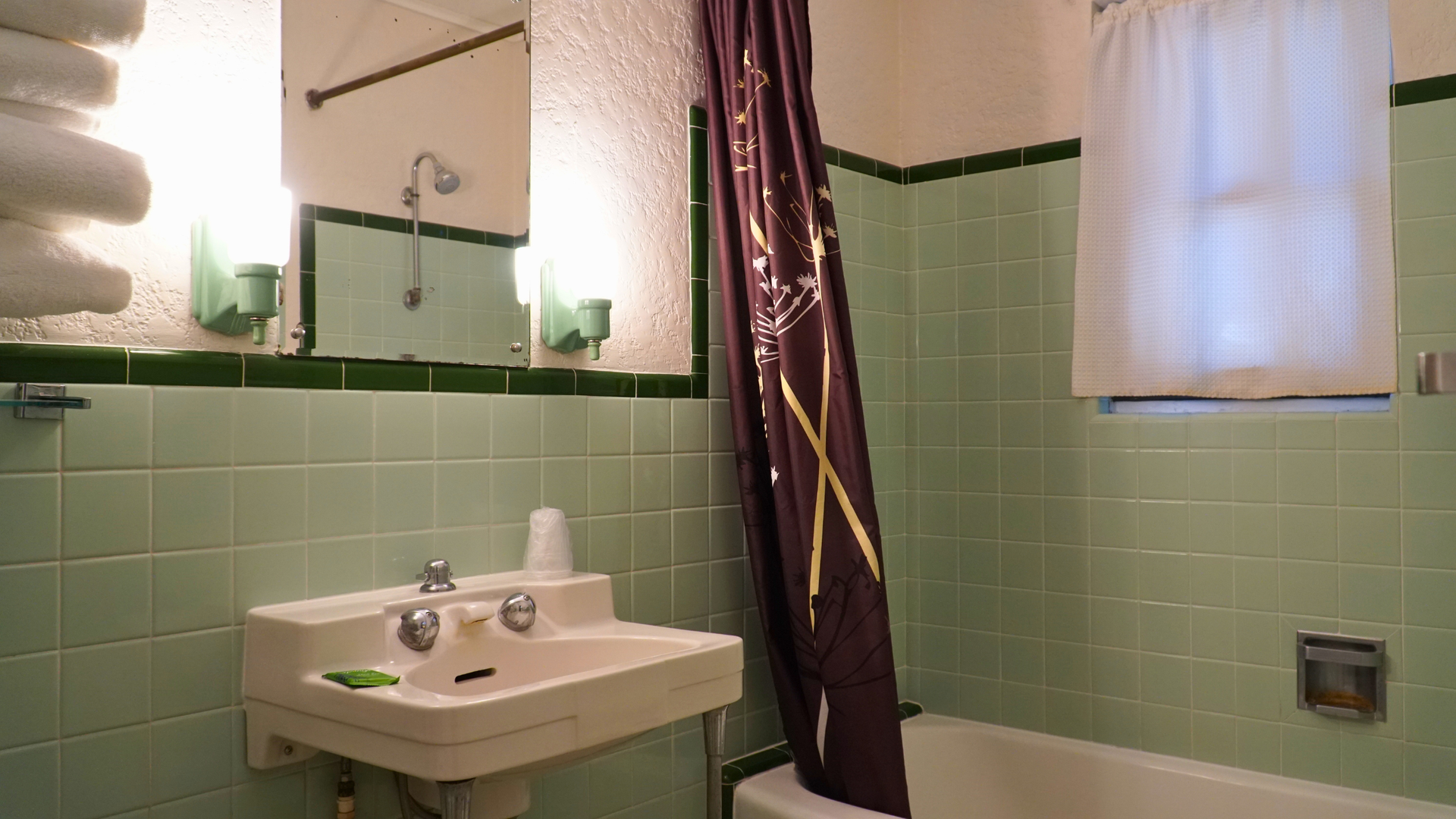 Small bathroom with green tile walls, pedestal sink, shower curtain, bathtub, and window.