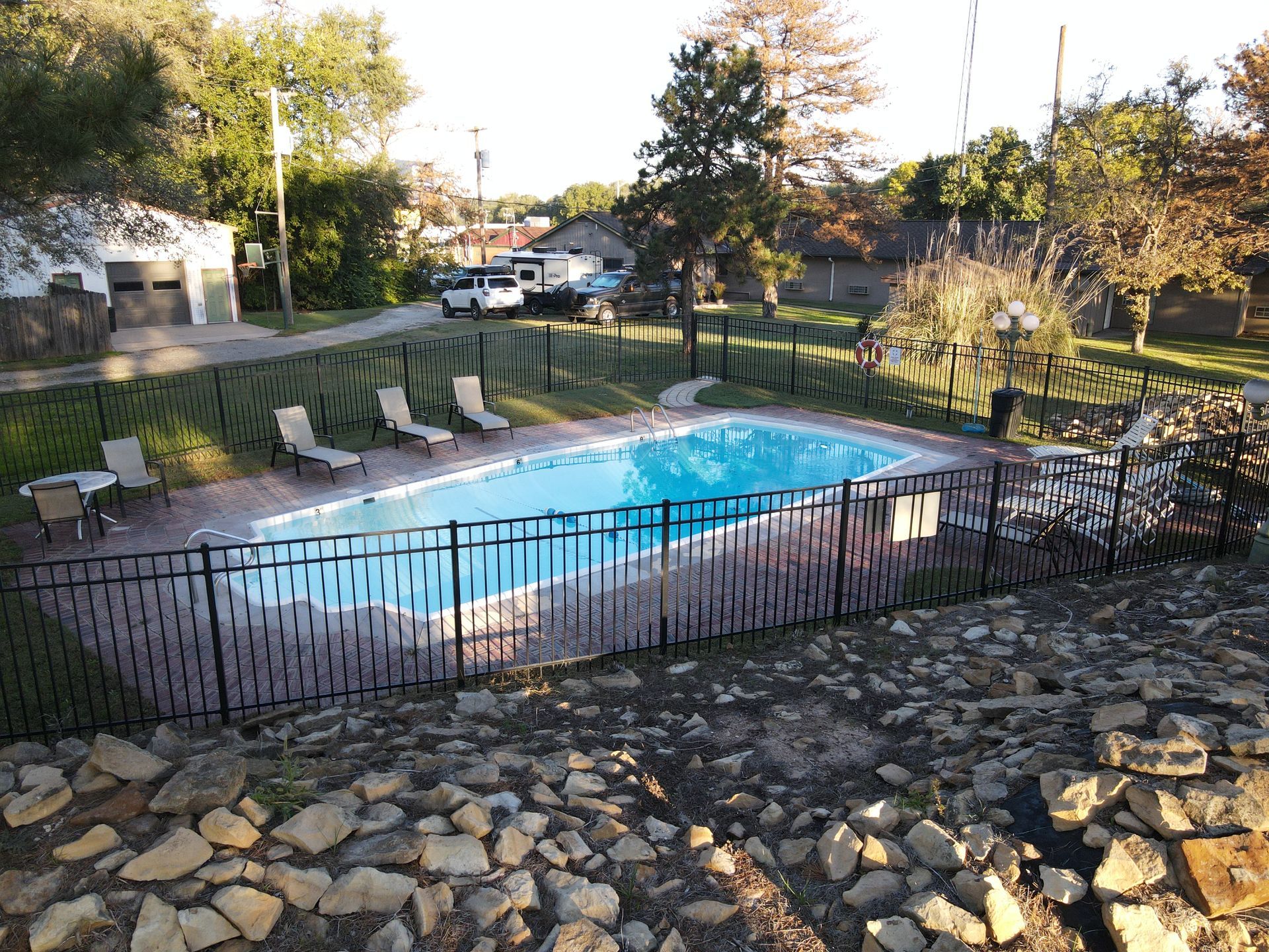 Outdoor swimming pool with lounge chairs behind a black fence, surrounded by trees and stone landscaping.
