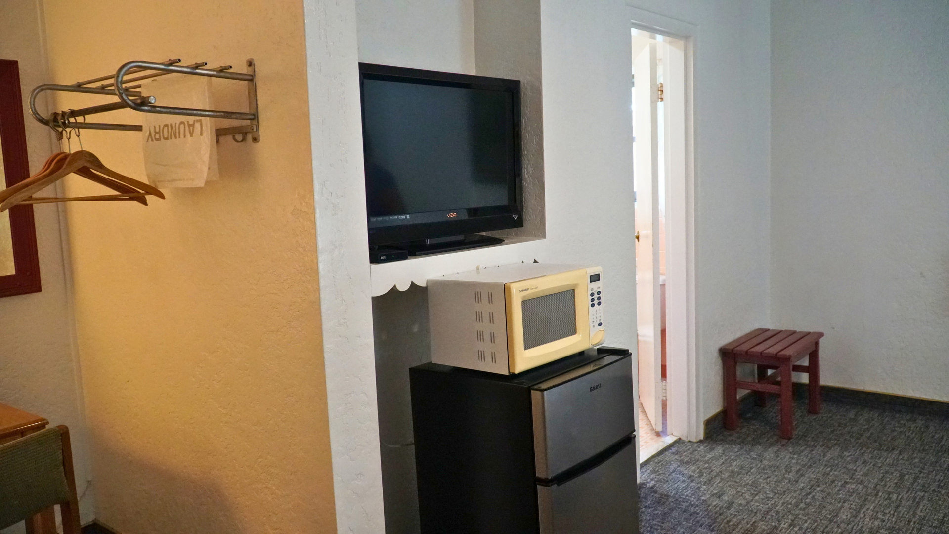 Small hotel room with wall-mounted TV, microwave on mini fridge, coat rack, and a stool near the doorway