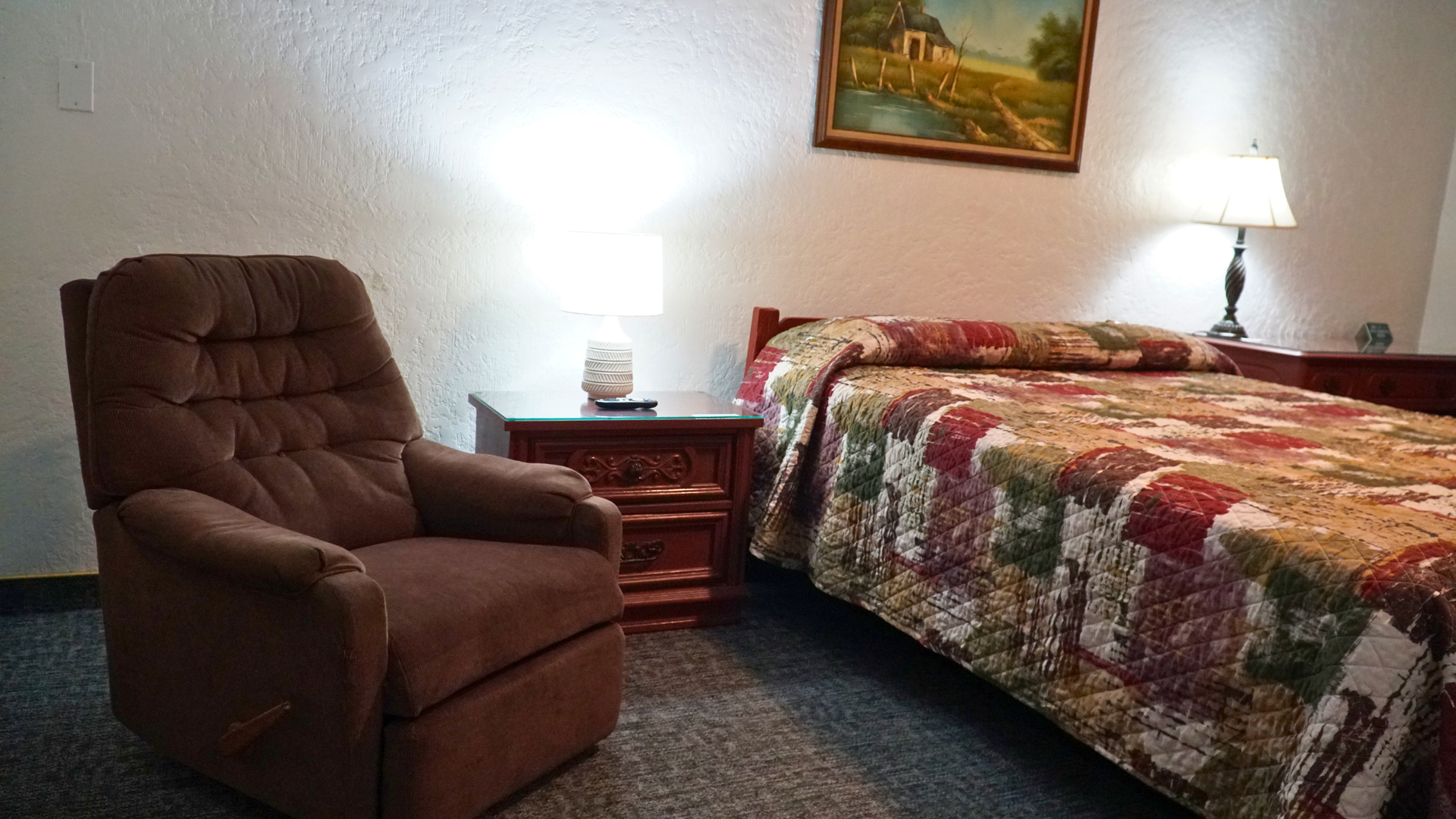 Cozy bedroom with a brown recliner, bedside table, lamp, and a colorful quilted bedspread.