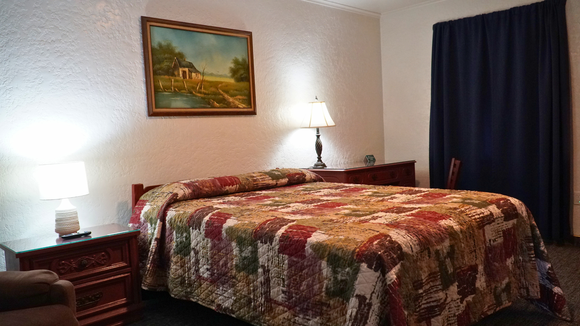 Bedroom with a patterned bedspread, two bedside lamps, a framed landscape, and dark curtains.