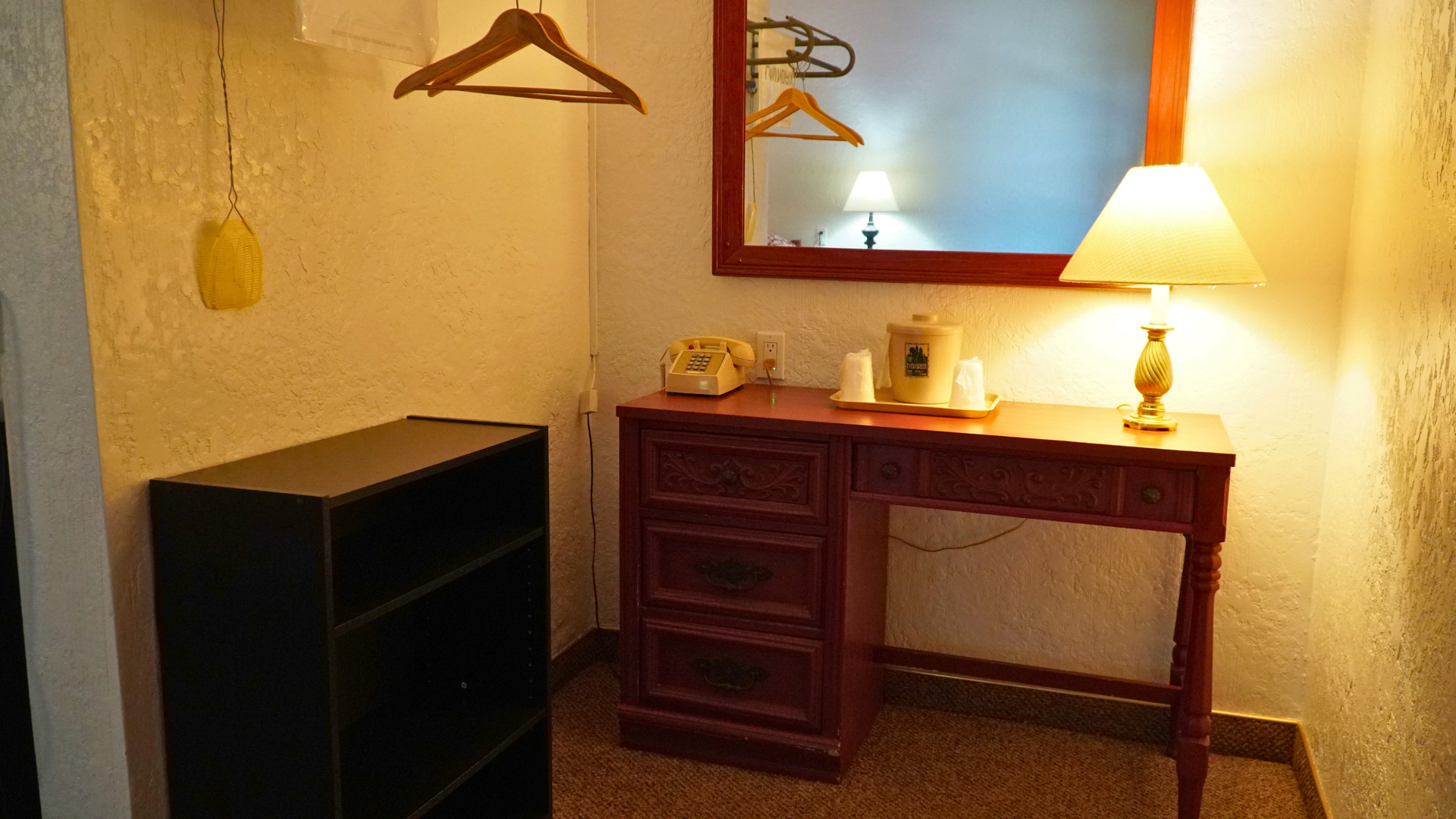 Small hotel room corner with desk, lamp, hanging clothes, and luggage rack beside a dresser.