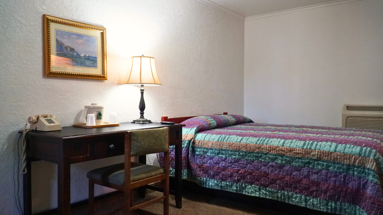 Small bedroom with a made bed, desk, lamp, and framed wall art.