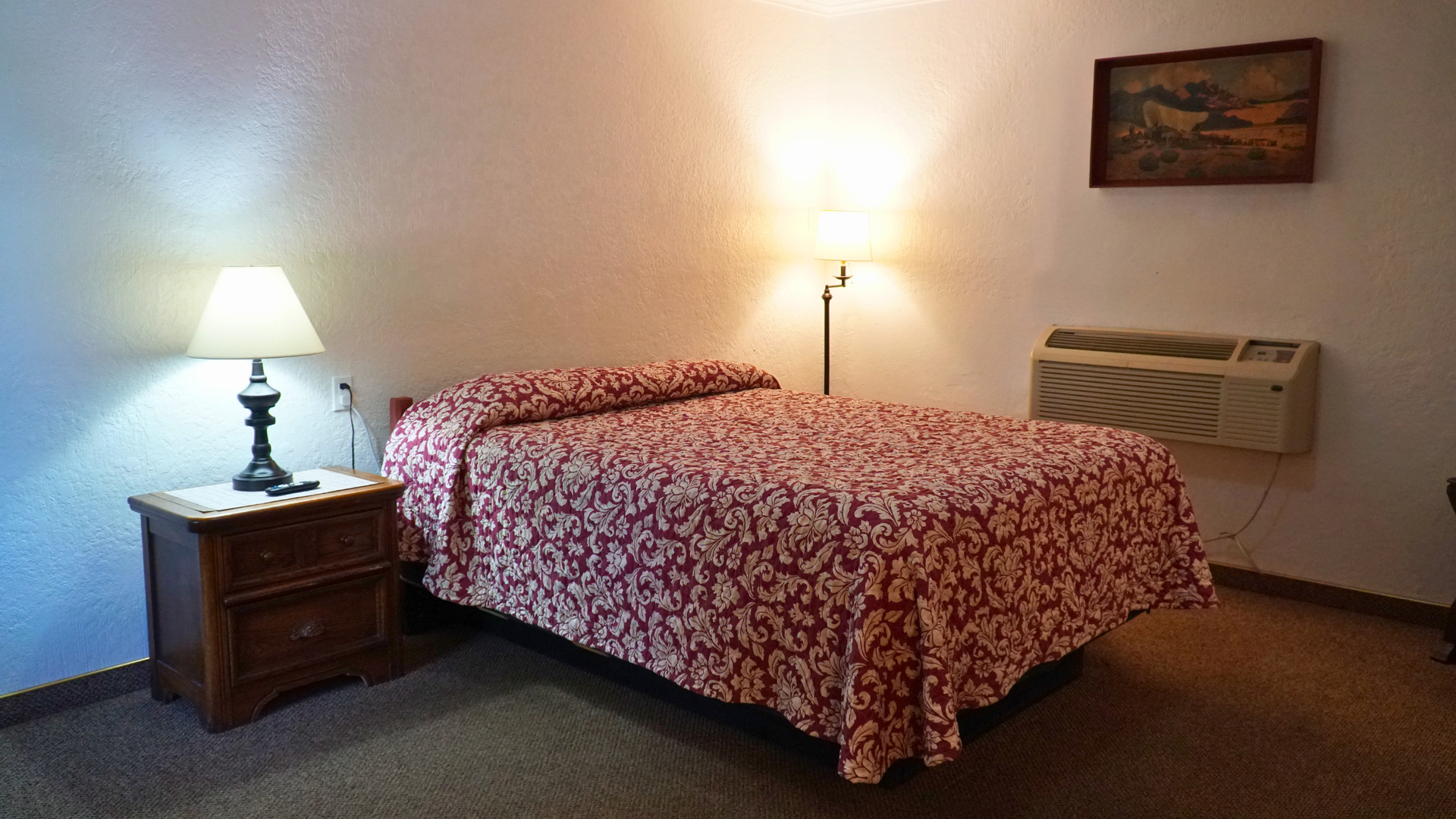 Cozy bedroom with patterned bedspread, bedside lamp, nightstand, wall art, and a wall heater