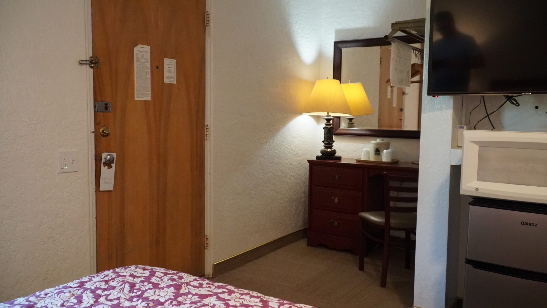 Small hotel room with bed, wooden door, dresser with lamp, mirror, and TV