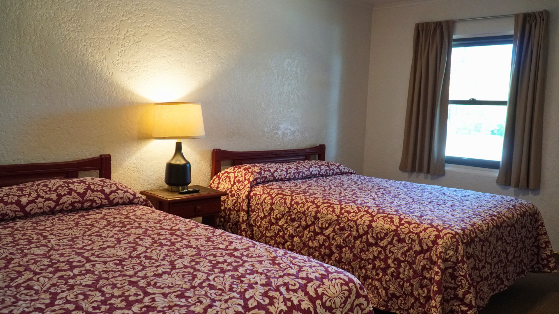 Bedroom with two patterned beds, a lamp on a nightstand, and a window with curtains
