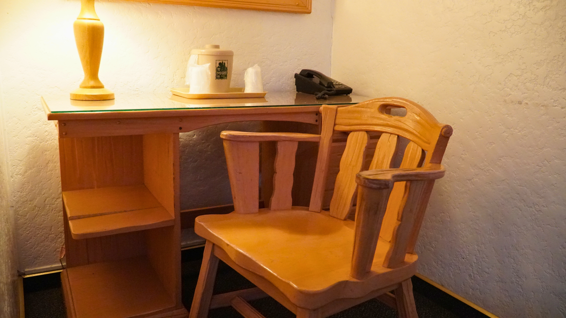 Wooden desk with chair, lamp, and phone in a warm-lit room corner.
