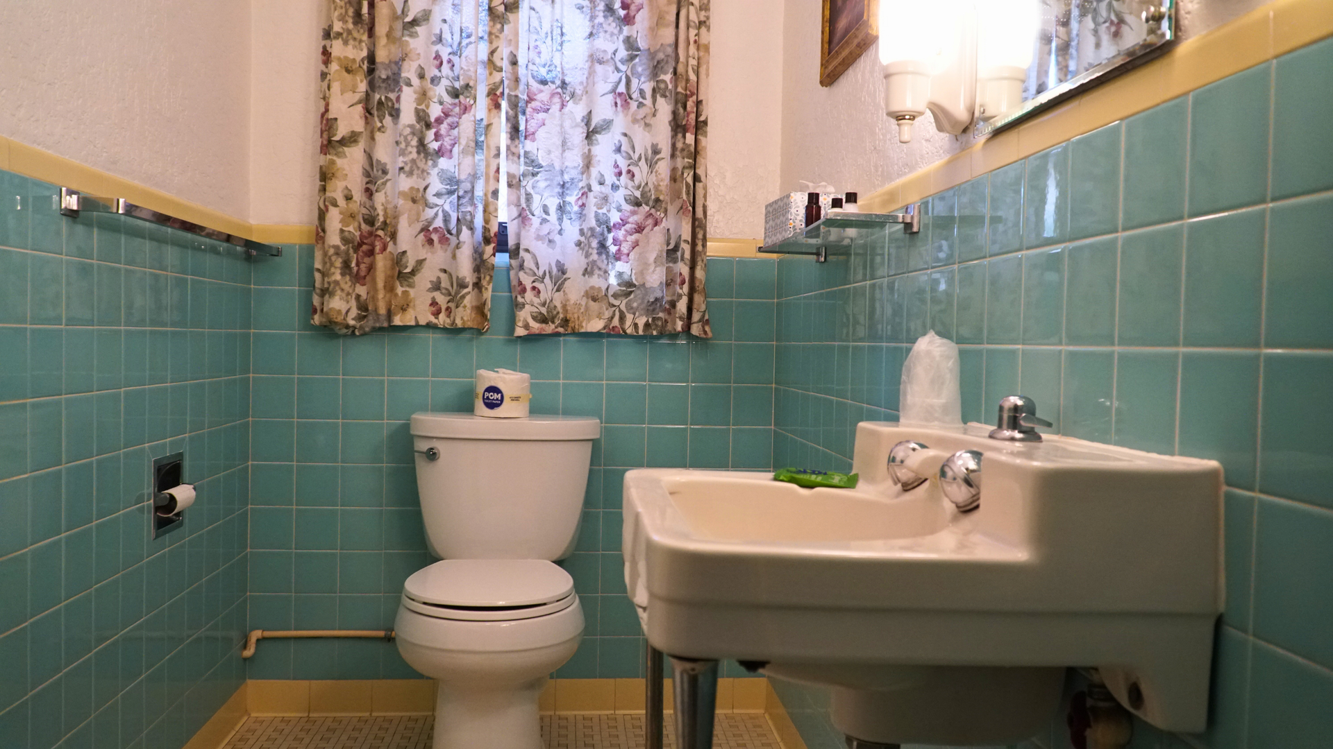 Small bathroom with turquoise tile, toilet, sink, and a floral curtain by the window.
