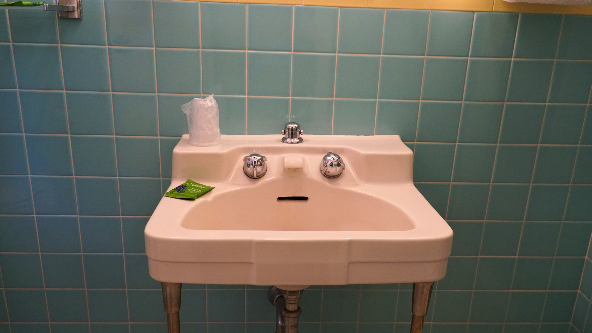 Small white sink with twin faucets against teal tiled bathroom wall, with a green cloth on the left side