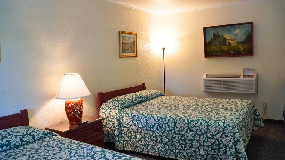 Small motel room with two beds, bedside lamp, wall art, and a window air conditioner.