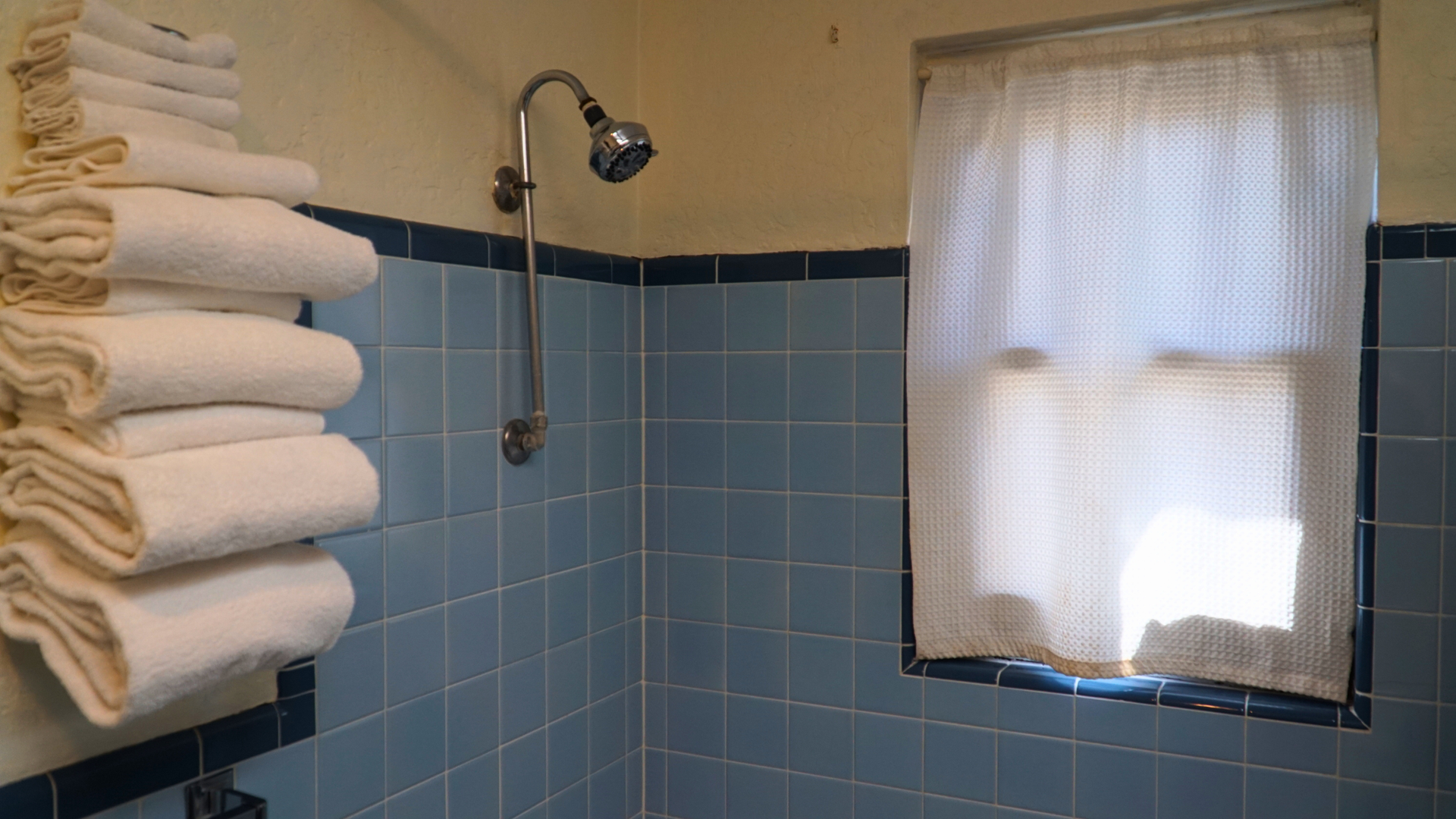 Small blue-tiled bathroom with stacked white towels, a showerhead, and a curtained window.