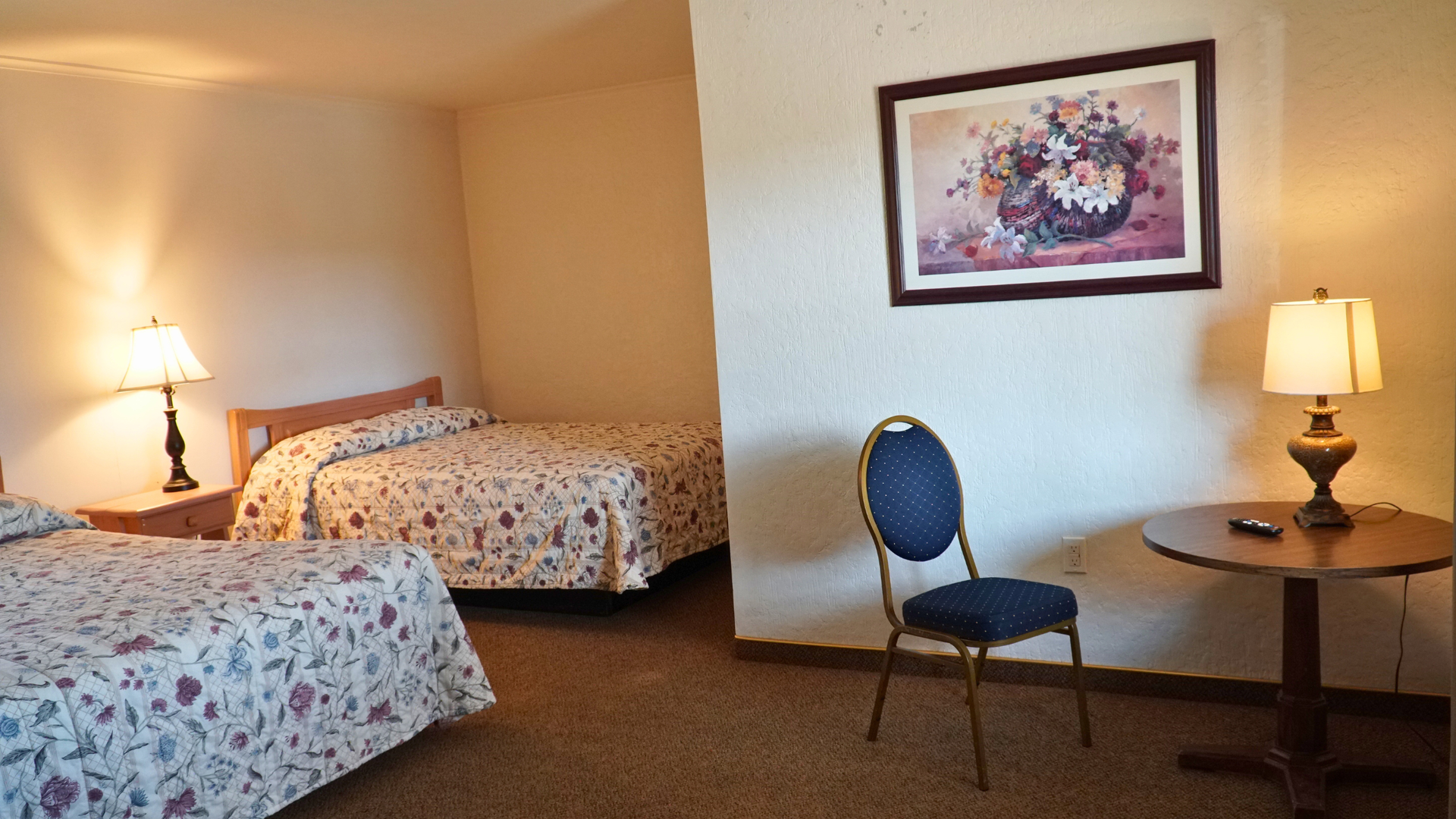 Small hotel room with two floral beds, lamps, a chair, table, and framed wall art.