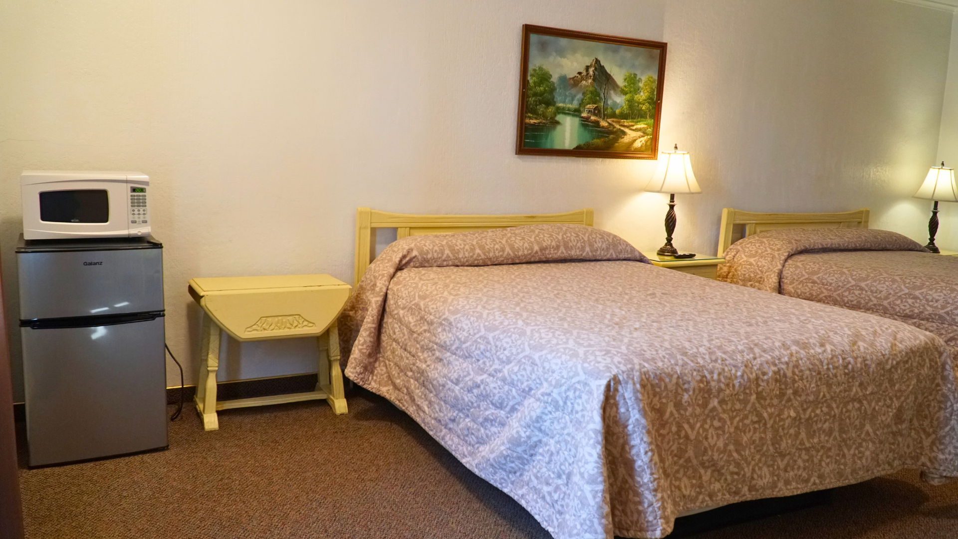 Hotel room with two beds, mini fridge and microwave, bedside lamp, and framed landscape painting