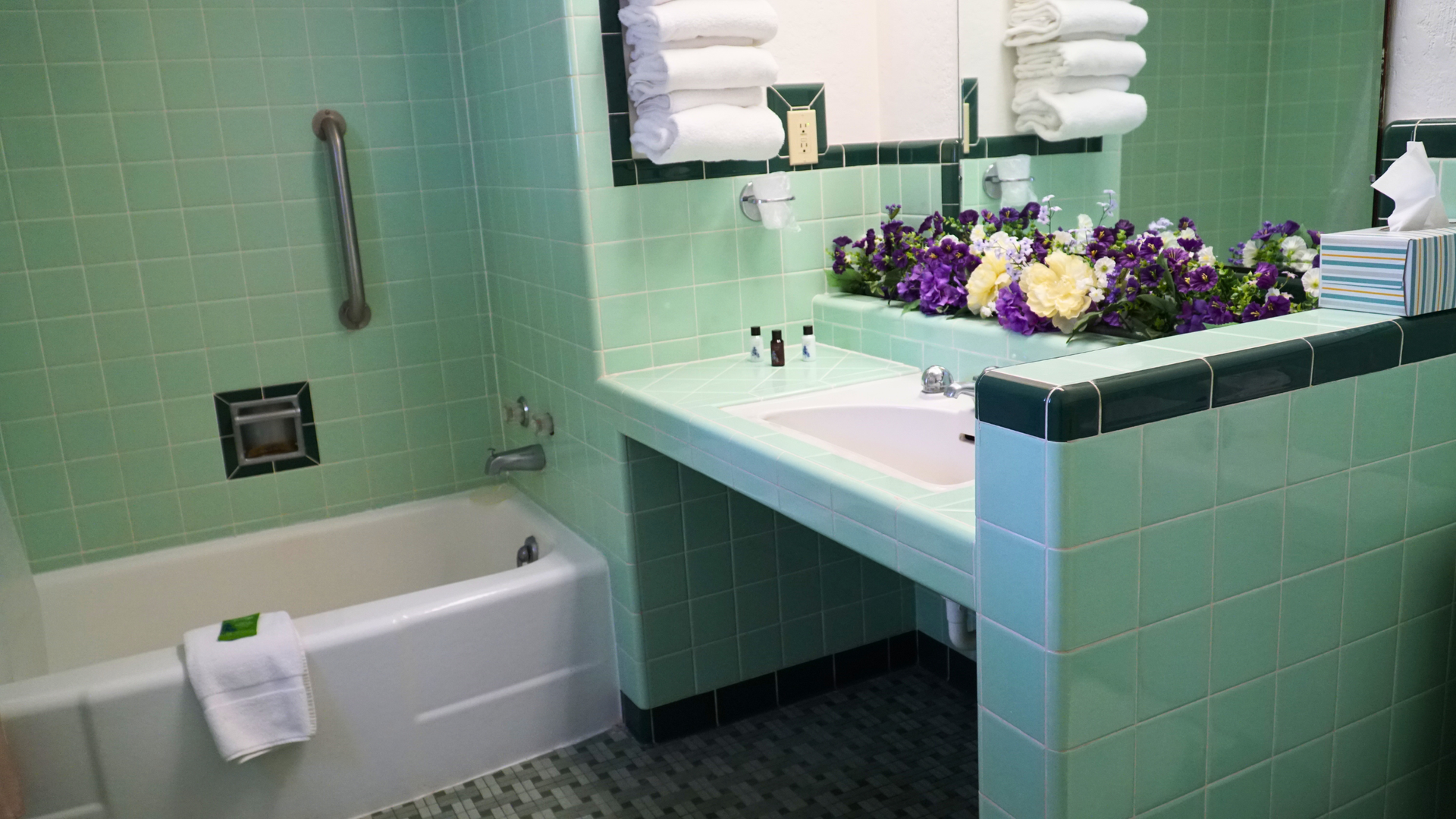 Bathroom with mint-green tile, bathtub, and vanity with sink, mirror, towels, and flowers.