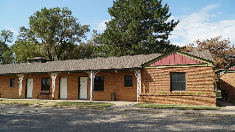 Single-story brick building with white doors and red trim, set among trees and a gravel lot.