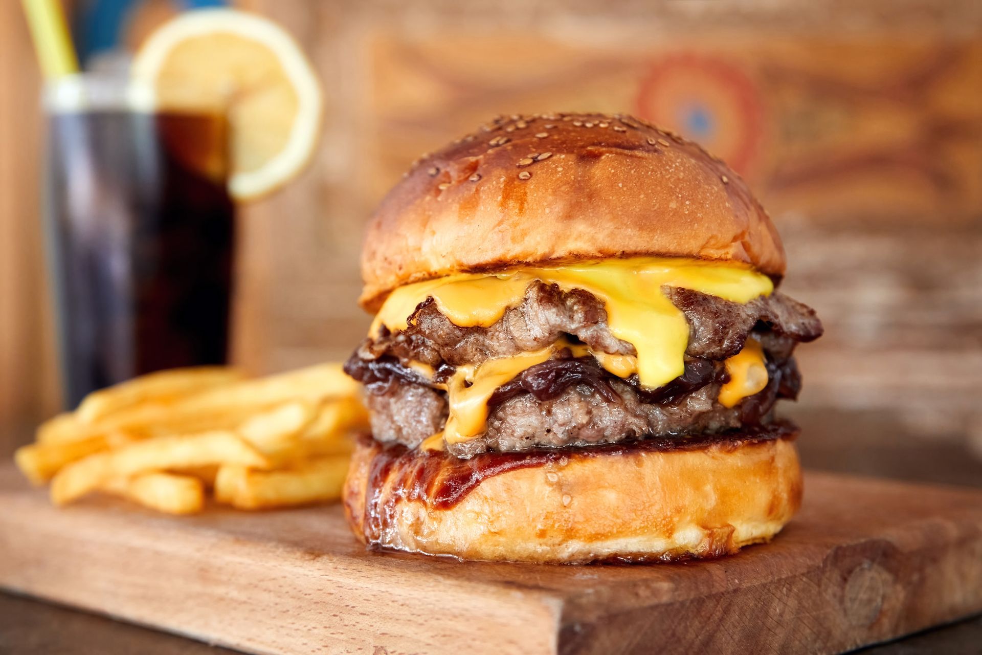 Cheeseburger with melted cheese on a wooden board, with fries and a drink in the background