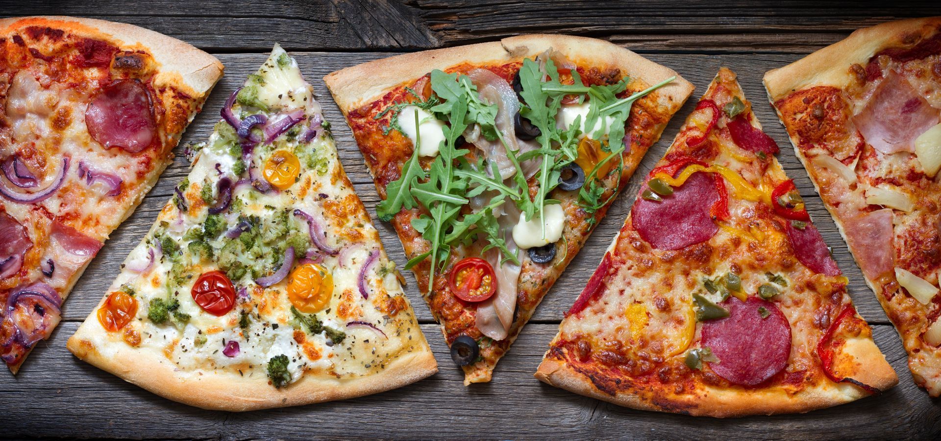 Assorted pizza slices with varied toppings arranged on a dark wooden surface.