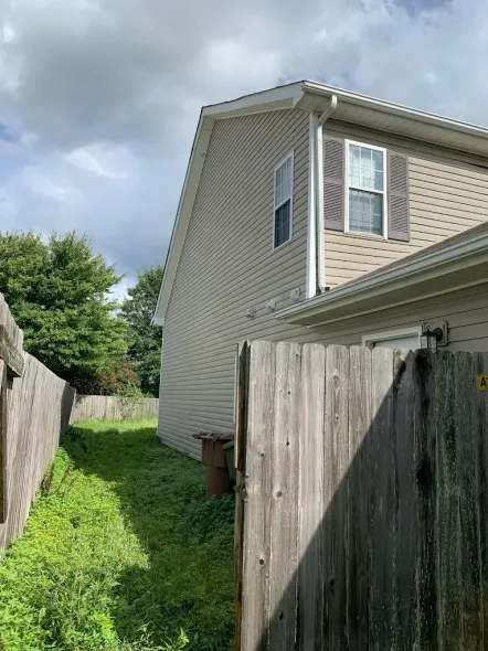 A narrow, grassy side yard between a tan house with vinyl siding and a wooden privacy fence under a cloudy sky.
